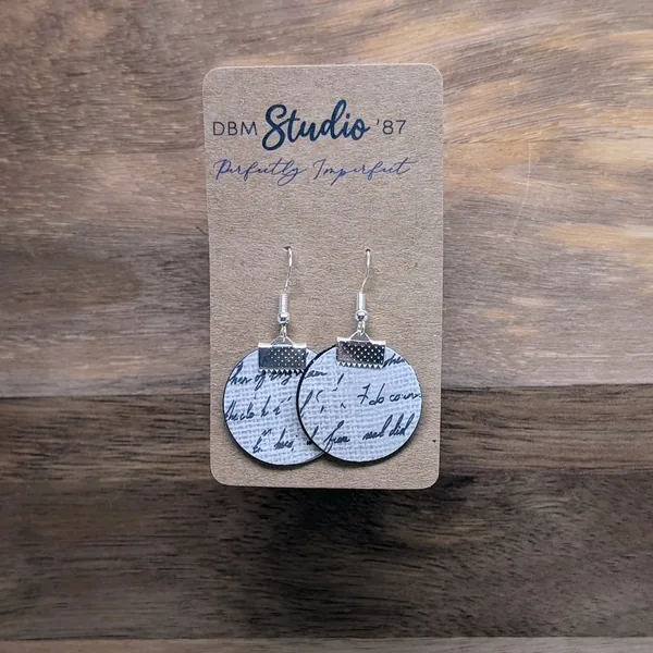 quarter size round earrings made from layers of paper modpodged together, white  with black cursive front and black with white cursive back. The color of the sides made by the paper inside is black. Silver-tone findings.